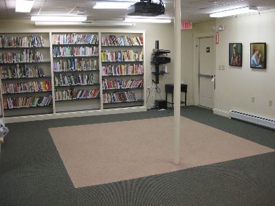 A small library room with shelves full of books along the walls, a large carpeted area in the center, and a single support pole. Two framed pictures hang on the wall near a door and an equipment stand.
