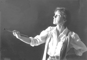 A person wearing a white shirt and vest stands in profile against a dark background, looking upward while holding a paintbrush in one hand and resting the other hand on their hip.
