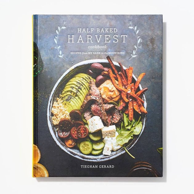 A cookbook titled Half Baked Harvest by Tieghan Gerard, featuring a bowl of salad with avocado, steak, feta cheese, carrots, and olives on the cover. The background is white and the book is viewed from above.