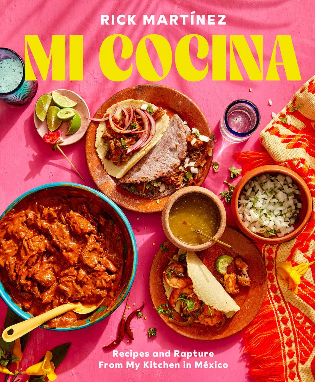 Overhead view of vibrant Mexican dishes on a pink table: a bowl of stew, tacos, lime slices, chopped onions, cilantro, a blue drink, and a yellow-patterned napkin. Text reads, MI COCINA by Rick Martínez.