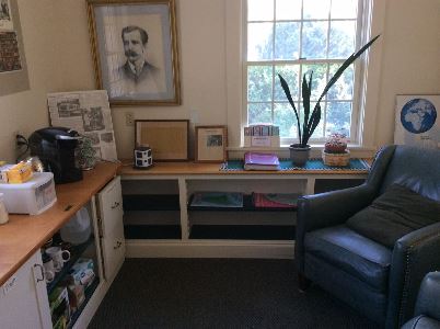 A cozy office corner with a framed portrait on the wall, a large window, a coffee maker on the counter, empty shelves, potted plants, and a cushioned armchair. Sunlight streams in through the window.