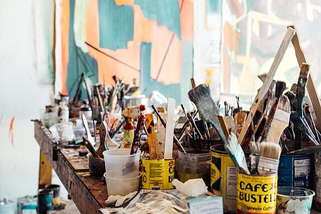 A cluttered artist’s workspace with jars and cans filled with paintbrushes, plastic cups, and art supplies on a wooden table; colorful abstract paintings hang in the background.