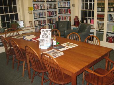 A wooden table with chairs around it is set in a library, with bookshelves, armchairs, and large windows in the background. Papers, books, pens, and a 1040 tax form booklet are on the table.